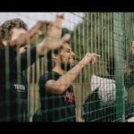 Three individuals are standing behind a green metal fence, smiling and gesturing with their hands through the gaps of the fence. The scene suggests they are likely enjoying an outdoor 5-a-side football game, with blurred greenery in the background. © Powerleague