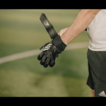 A person wearing a white shirt and black shorts is adjusting a black and white goalkeeper glove on their left hand. The blurred sports field in the background suggests a 5-a-side football environment. The person's face is not visible. © Powerleague