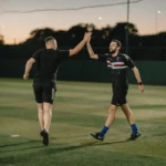 Two men on a soccer field, high-fiving each other. One is jumping in the air. Both are wearing black athletic gear. The field is outdoors, and it appears to be dusk with a darkening sky in the background. The scene conveys a sense of achievement and camaraderie, reminiscent of veterans celebrating a football victory. © Powerleague