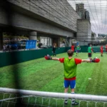 A soccer game is in progress on an urban rooftop field. A goalkeeper wearing a green bib and red gloves stands with arms outstretched in front of the goal. Other players, also in colorful bibs, are scattered across the green artificial turf—this dynamic setting is perfect for team building days. © Powerleague