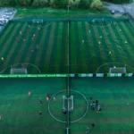 Aerial view of two adjacent fenced soccer fields with players actively engaged in matches. Representing local football leagues, teams are distinguished by red and yellow kits on the left field, and dark and neon green kits on the right field. Surrounding areas feature trees and parked cars. © Powerleague