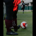 A soccer player in black uniform and maroon socks stands next to a soccer ball on the field, holding a red training bib in hand. Another person in the background, also wearing a black uniform, is walking away. The scene appears to be before a match or training session in one of the mixed football leagues. © Powerleague