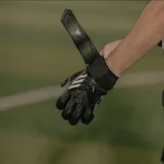 A person wearing black soccer goalkeeper gloves is tightening the strap on one glove with their other hand. The gloves boast a sleek design with white accents. In the background, possibly at the home of 5-a-side, the scene is blurred, hinting at an intense soccer field setting. © Powerleague