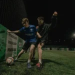 Two young men are playing soccer at night on an outdoor field. The player in the blue jersey is trying to shield the ball from an opponent in a black jersey. The field, surrounded by fences and lit by artificial lighting, is adorned with Powerleague banners showcasing advertising and partnerships. © Powerleague