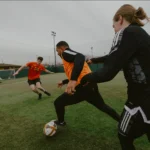 Three soccer players are in action on a green field. Two players in black and orange jerseys move toward the ball, while a fourth player in a red jersey runs towards them from the background. The scene appears competitive and dynamic, reminiscent of intense moments seen in football tournaments. © Powerleague