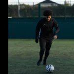 A person with curly hair, dressed in black athletic wear and black sneakers, is playing soccer on a fenced outdoor field. Under the watchful eyes of 5-a-side referees, they are in motion, appearing focused as they approach a soccer ball on the ground. The background includes trees and a building. © Powerleague