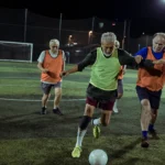 A group of older men playing walking football on an outdoor field at night. One man in a green vest is actively dribbling the ball while others, dressed in orange vests and casual sportswear, move around the field. The background features a goalpost and netting. © Powerleague