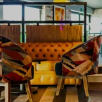 A cozy remote workspace featuring a yellow tufted leather couch and two patterned upholstered chairs. The chairs have a colorful geometric design, and the space includes wood flooring with large windows in the background, allowing natural light to permeate the room. © Powerleague