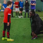 A young boy in a red and black soccer uniform stands on grass, talking to a coach who is kneeling and tying his own shoe. In the background, slightly out of focus, other children in various uniforms engage in junior football training on the soccer field. © Powerleague