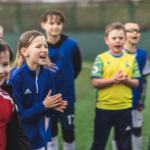 A group of young children stands on a soccer field, smiling and clapping. They are wearing various soccer jerseys and appear to be celebrating or cheering at the Powerleague Bradford opening event. The background shows a green fence and a slightly blurred landscape, indicating an outdoor setting. © Powerleague