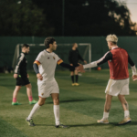 Two soccer players in white uniforms give each other a high-five on an outdoor field during dusk. In the background, other players and goalposts are visible. One player wears a red training vest, and the scene is lit by twilight. © Powerleague