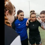 A group of six people stands closely together in a huddle on a sports field. They have their arms around each other’s shoulders, displaying a team spirit. They are dressed in athletic wear and appear focused and determined. © Powerleague