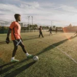 A soccer player in an orange jersey and black shorts stands on a field, poised to kick a soccer ball. Several other players are visible in the background under a clear sky with the sun setting, casting long shadows on the field. © Powerleague