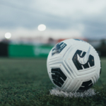 Close-up of a soccer ball with a black and white pattern resting on artificial turf. The background is out of focus, showing a sports field with goalposts and blurred lights in the distance, suggesting an evening or early morning setting. © Powerleague