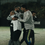 A group of four men wearing athletic clothing, including jerseys and shorts, are seen celebrating and hugging on an outdoor soccer field at night. The mood is festive, indicating they managed to stoke their camaraderie to excel in this moment of triumph. © Powerleague