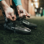 A person wearing a teal long-sleeve shirt is bent down tying the laces of black and white cleats with a noticeable Nike logo. The setting appears to be an indoor sports facility with a turf floor. © Powerleague