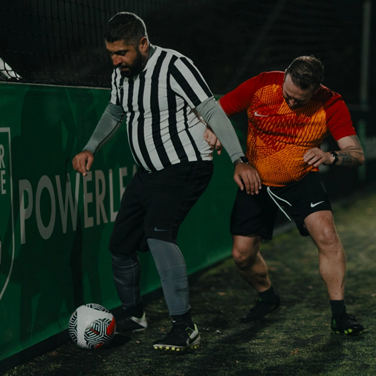 Two men are playing soccer on an outdoor field at night. One man, wearing a black and white striped referee jersey, is dribbling the ball near the sideline. The other man, in an orange and red jersey, is closely defending him. A green fence and 