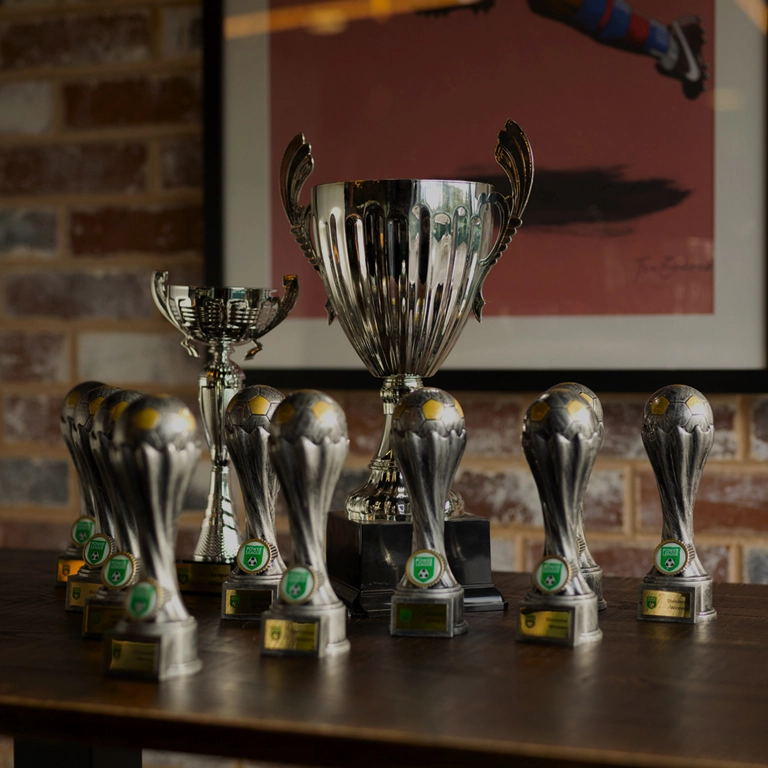 A collection of silver trophies displayed on a wooden table. The centerpiece is a large, two-handled trophy at the back, flanked by a smaller, ornate cup and several smaller soccer ball-shaped trophies lined up in front. The background features a framed picture. © Powerleague