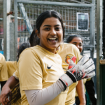 A young woman wearing a yellow soccer jersey and goalie gloves smiles widely at the camera. She stands in front of a metal fence, with other teammates in similar jerseys visible in the background. The scene portrays a joyful moment during a women's football event. © Powerleague