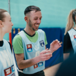 Three people, wearing sports bibs with sponsorship logos, smile and laugh together, standing in what appears to be an indoor sports venue with a net visible in the background. © Powerleague