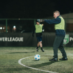 A person wearing a green bib and gloves is on an outdoor football field at night, playing as a goalkeeper. There's a blue and white football in front of them. Another player is in the background. The field is surrounded by a fence with 