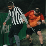 Two men are playing soccer on an outdoor field. The man on the left, in a black-and-white striped shirt, controls the ball while closely guarded by the man on the right, wearing a red-orange shirt. It's nighttime, with a dark fence and green advertisement board in the background. © Powerleague