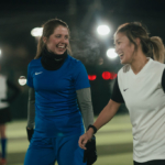 Two women, one in a blue soccer uniform and the other in a white and black uniform, are smiling and walking together on a lit outdoor soccer field at night. Both are wearing gloves, and there are other players and lights visible in the background. © Powerleague