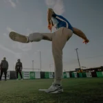 A person in athletic wear, mid-kick, with one leg extended up and arms slightly out for balance, on a sports field. Two others stand in the background near a fence under a clear sky, possibly waiting or watching. Join a small-sided football league at Powerleague to experience moments like these. © Powerleague