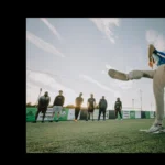 A person wearing a blue shirt and beige pants is shown mid-kick on a soccer field with an overcast sky. Five individuals stand watching in the background near the goal, wearing dark clothing. The image captures movement and action with a wide, clear sky backdrop—join a small-sided football league at Powerleague. © Powerleague