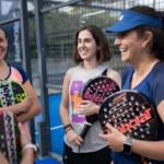 Four women are standing together on a padel court, holding their racquets and dressed in sportswear. They are all smiling and engaging in conversation, suggesting camaraderie and enjoyment. The background shows the fenced area and part of the court surface, highlighting their shared love for padel. © Powerleague