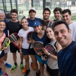 A group of eleven people is gathered for a selfie on a padel tennis court. They are smiling, dressed in athletic clothing, and holding paddles. The court is surrounded by a fence with glimpses of other courts and buildings in the background. © Powerleague