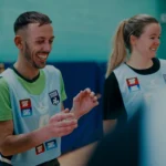 A man and a woman, both wearing light blue sports bibs, laugh and smile while seen indoors, possibly in a sports facility or gym. The man is gesturing with his hands, and both are wearing green shirts under their bibs bearing various logos. They appear ready for a friendly game of mixed-netball. © Powerleague