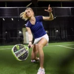 A woman wearing a blue t-shirt and white skirt lunges to hit a ball with a paddle racket on an indoor padel court. Bright lights illuminate the padel court, highlighting her athletic movement and focused expression. © Powerleague