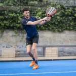 A man dressed in a navy blue shirt and black shorts is playing padel on a blue court. He is in mid-swing, holding a red racket with both hands. The background features a stone wall covered in green ivy, creating an ideal setting for an intense padel match. © Powerleague