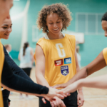 A group of women wearing yellow sports jerseys are standing in a circle, placing their hands together in the center. They appear to be a sports team, united and ready to participate. A basketball court is visible in the background. © Powerleague