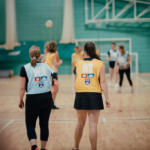 A group of netball players are on an indoor court. Two players in focus, wearing different color bibs, are standing with their backs to the camera, both watching the ball mid-air. The background includes other players, a hoop, and a green wall. © Powerleague