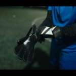 A soccer player wearing a blue uniform and black long sleeves is seen adjusting black gloves on a dimly lit field, almost like preparing for some legal stuff. Only the torso and arms are visible in the image. The background is out of focus. © Powerleague