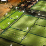 A nighttime aerial view shows multiple well-lit, fenced soccer fields with players engaging in games. Surrounding the fields are parking areas and streets with cars, illuminated by streetlights. The scene is vibrant with the activity of players and the glow of field lights. © Powerleague