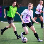 Two women are playing women's football on a green field. One in a green bib and black shorts is defending, while the other in a white and purple shirt and black shorts is dribbling the ball. Another player in the background is running towards them. © Powerleague