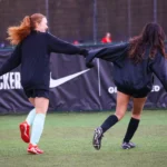 Two female soccer players on a field hold hands and laugh, running in opposite directions at the home of 5-a-side. They are wearing black jackets, with one in light turquoise socks and red cleats, and the other in black socks and black cleats. A banner with logos is visible in the background. © Powerleague