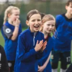 A group of children in sports attire are gathered on a soccer field, reminiscent of vibrant football holiday camps. The focus is on a girl in the center wearing a blue jacket, clapping and smiling with enthusiasm. Other children around her are also smiling and wearing various sports jerseys. © Powerleague