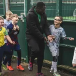 A group of children in colourful football uniforms joyfully follow a smiling coach on a damp, concrete path near a green fence. Most kids are laughing, and one boy on the right is playfully interacting with the coach. The weather appears cool and overcast, perfect for this football birthday party. © Powerleague