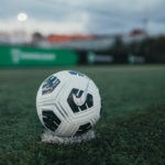 A close-up of a white and black soccer ball on a green artificial turf field, resting on a small patch of white marking. In the blurred background, there are lights on poles and a green banner, indicating an outdoor sports facility—perhaps where careers at Powerleague begin.