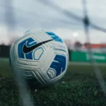 A close-up shot of a white and blue Nike soccer ball on a green grass field, positioned near the goal net. The blurred background reveals part of the goal post and a distant view of the sports facility, which also offers function room hire, under an overcast sky. © Powerleague