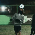 Two people play soccer at night on a field. The player in focus, wearing a gray hoodie and black shorts, skillfully balances a soccer ball on his head. The other player, slightly blurred, watches. A green fence and illuminated background hint at local football leagues in action. © Powerleague