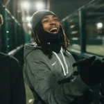 A person wearing a grey beanie, dark gloves, and a grey jacket is smiling and laughing in an outdoor setting at night. The background is dimly lit, featuring tall mesh fencing and bright lights from the local football leagues nearby. © Powerleague