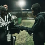 Three individuals are on a dimly lit sports field at night. Two are wearing dark hoodies and winter hats; one is opening a black Nike bag while the other looks on. The third person, likely a student, stands in the background in a black and white striped jersey, preparing for their late-night 5-a-side game. © Powerleague