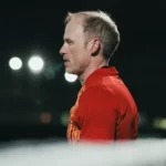 Side profile of a man with short, light hair standing outdoors at night. He is wearing a red shirt and appears focused, much like one of the 5-a-side referees. The background is dimly lit with a few bright lights visible in the distance. © Powerleague