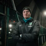 A person in a black beanie and jacket stands with arms crossed on a dimly lit soccer field. The individual has a focused expression and wears a green emblem with a soccer ball and 