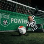 A soccer player in a black and white striped jersey dives to the ground in front of a goal, attempting to block a ball that is flying towards the net. A green Powerleague banner is displayed in the background, hinting at one of the local football leagues. The scene appears to be set at night.
 © Powerleague